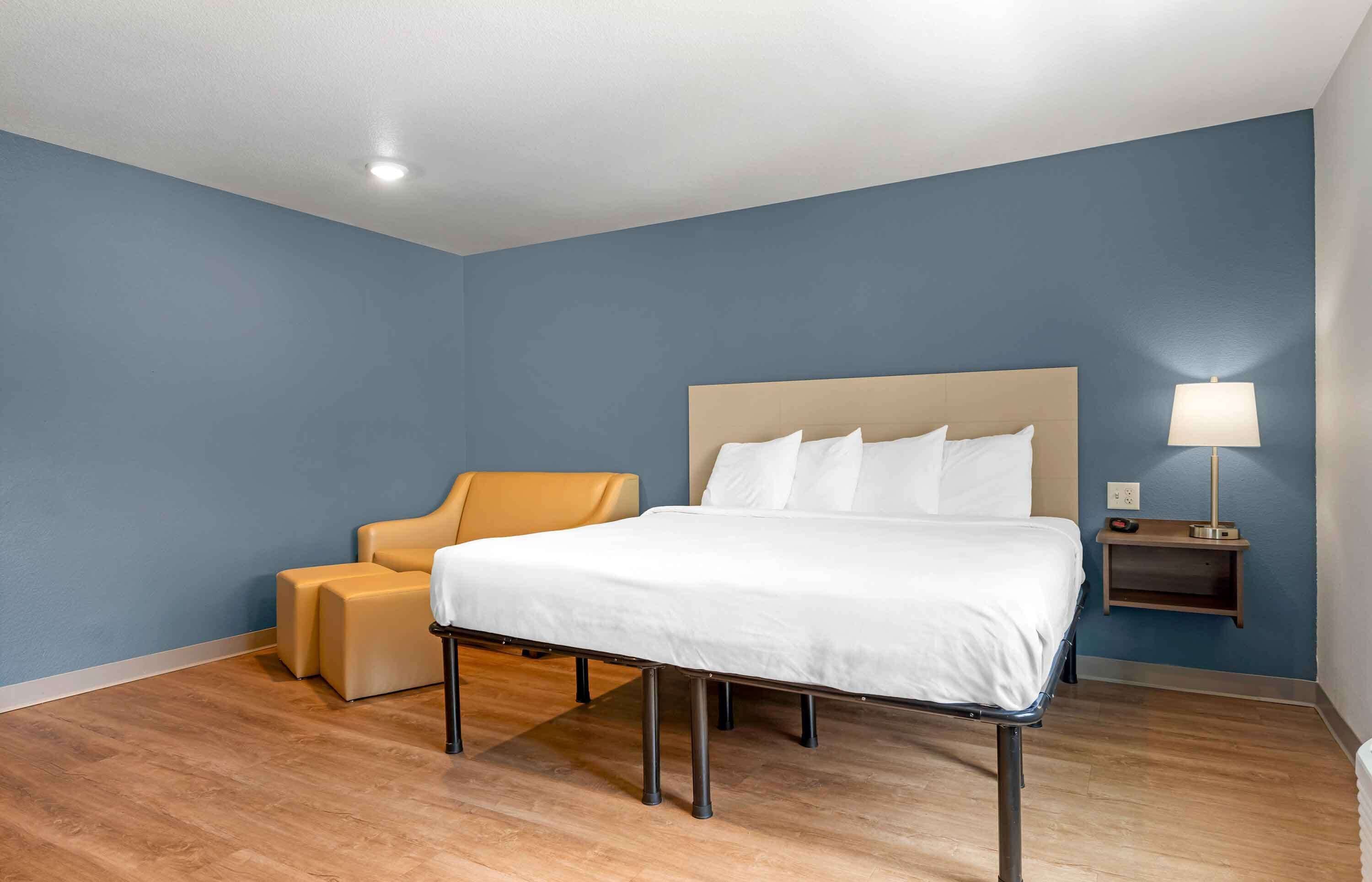 A hotel room with a bed, a chair, a nightstand with a lamp, and a wall-mounted shelf.