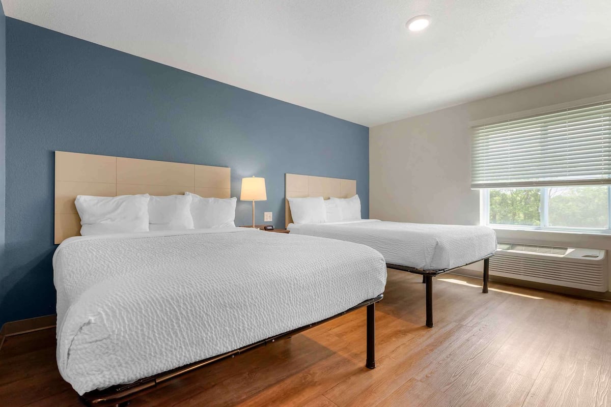 A hotel room with two beds, a nightstand with a lamp, a window with blinds, and a built-in air conditioning unit.