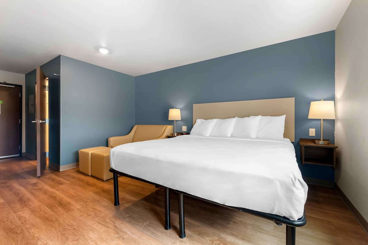 A modern hotel room with a large bed, two bedside tables with lamps, a sofa, and a wooden floor.
