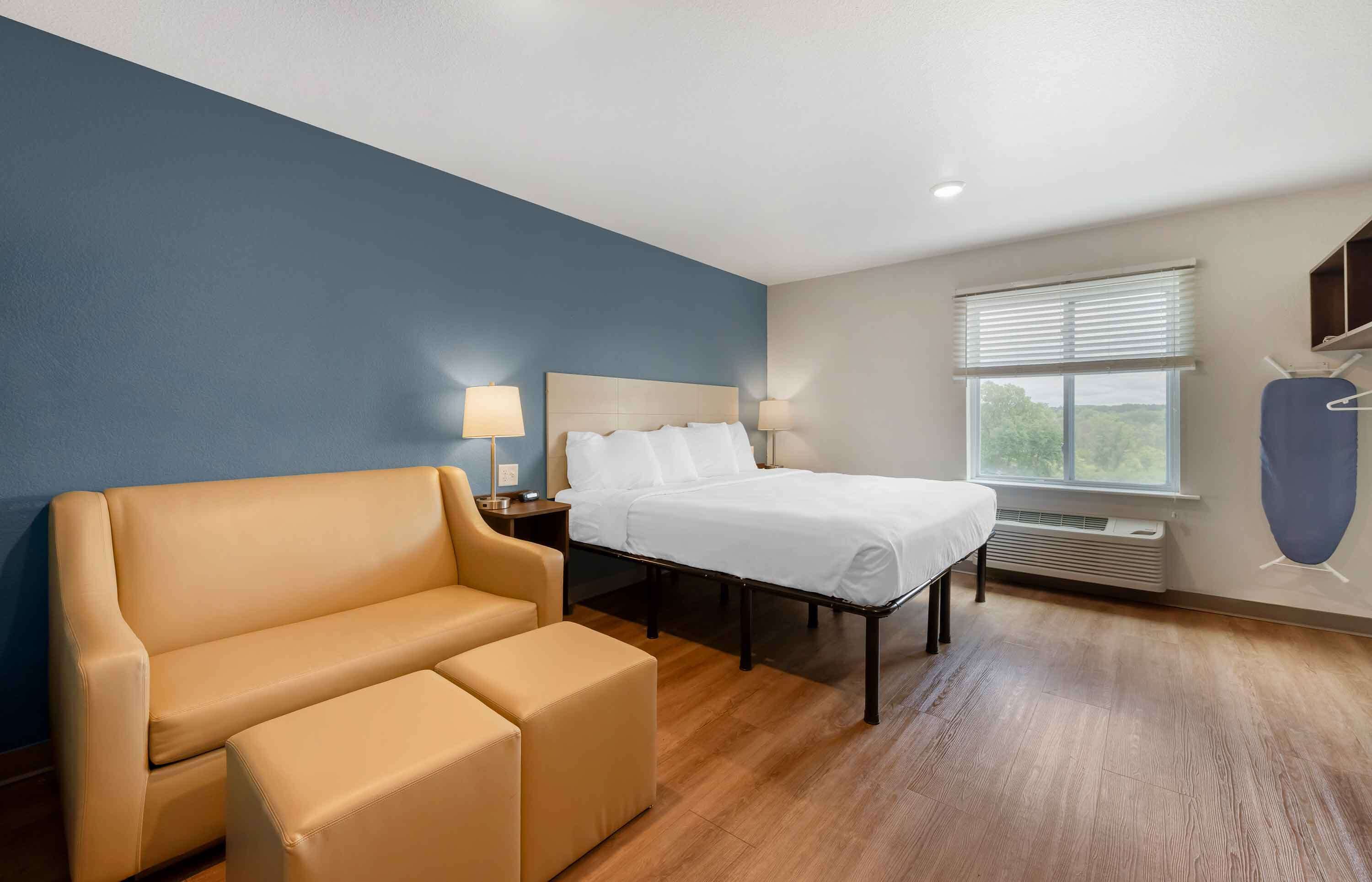 A hotel room with a bed, a sofa, a nightstand with lamps, a window with blinds, and a wall-mounted air conditioning unit.