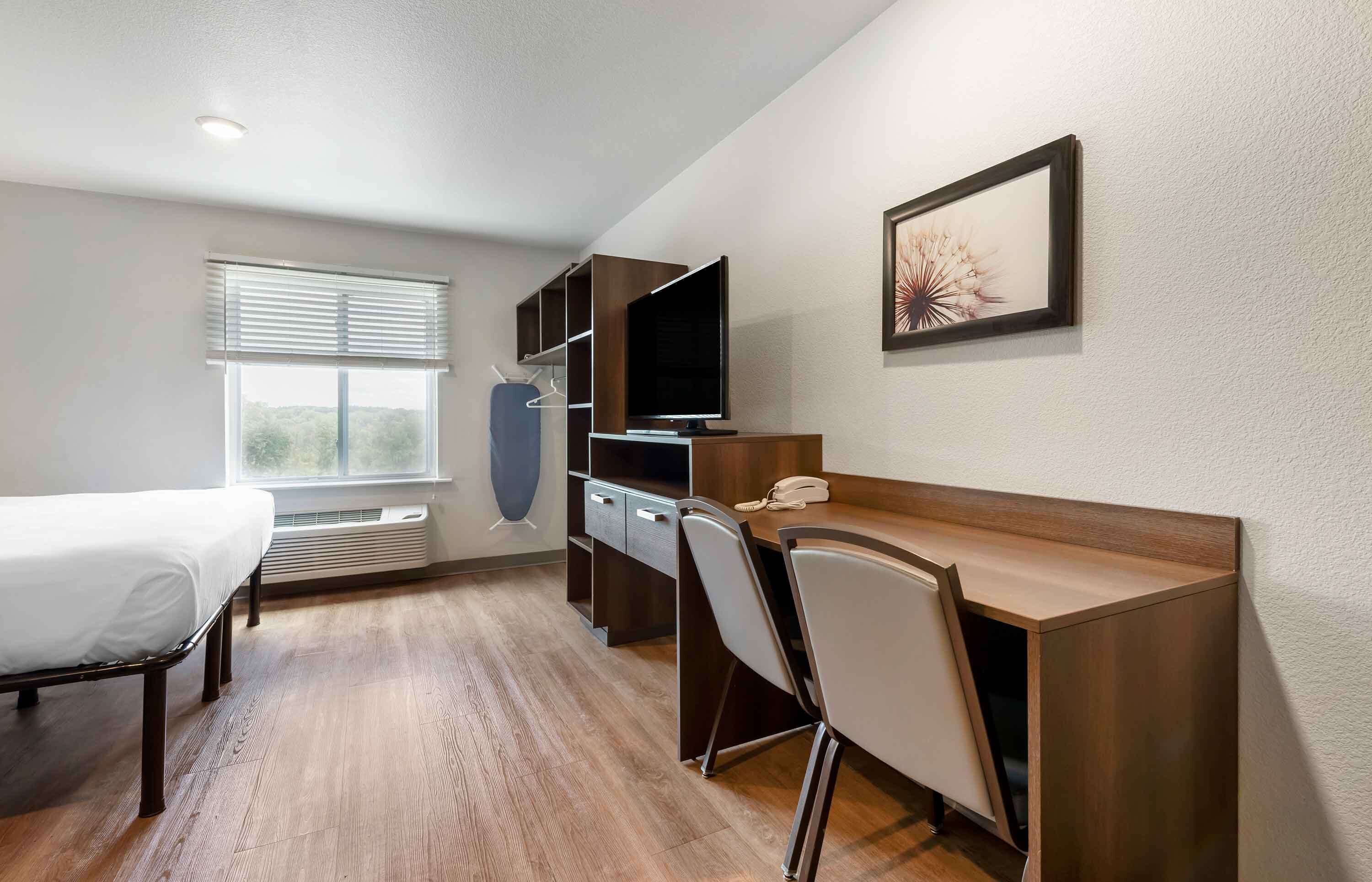A modern hotel room with a wooden desk, a chair, a bed, a TV, and a window with blinds.