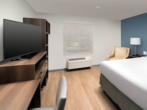 A hotel room with a flat-screen TV, a chair, a bed, a lamp, and a window with blinds.