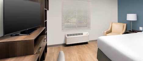A hotel room with a flat-screen TV, a chair, a bed, a lamp, and a window with blinds.