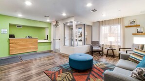 A hotel lobby with a reception desk, a blue ottoman, a sofa with patterned cushions, and a patterned rug. - WoodSpring Suites Harrisburg Linglestown (Harrisburg)