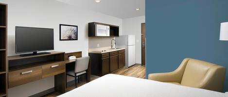 Quarto de hotel moderno com cama, escrivaninha com computador, cadeira, televisão, área de cozinha com micro-ondas e frigorífico, e uma parede de destaque azul.