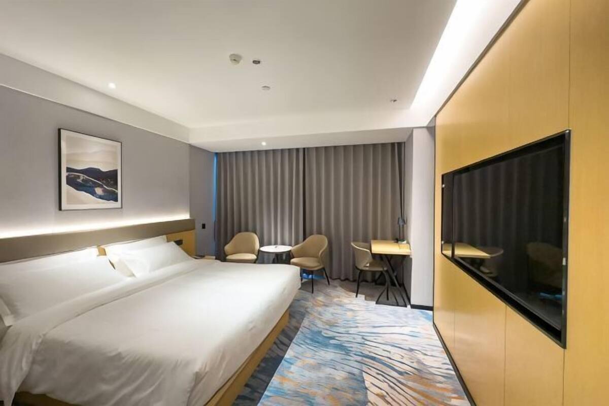 Ett hotellrum med en stor säng, två stolar, ett litet bord, en tv och ett stort fönster med gardiner.