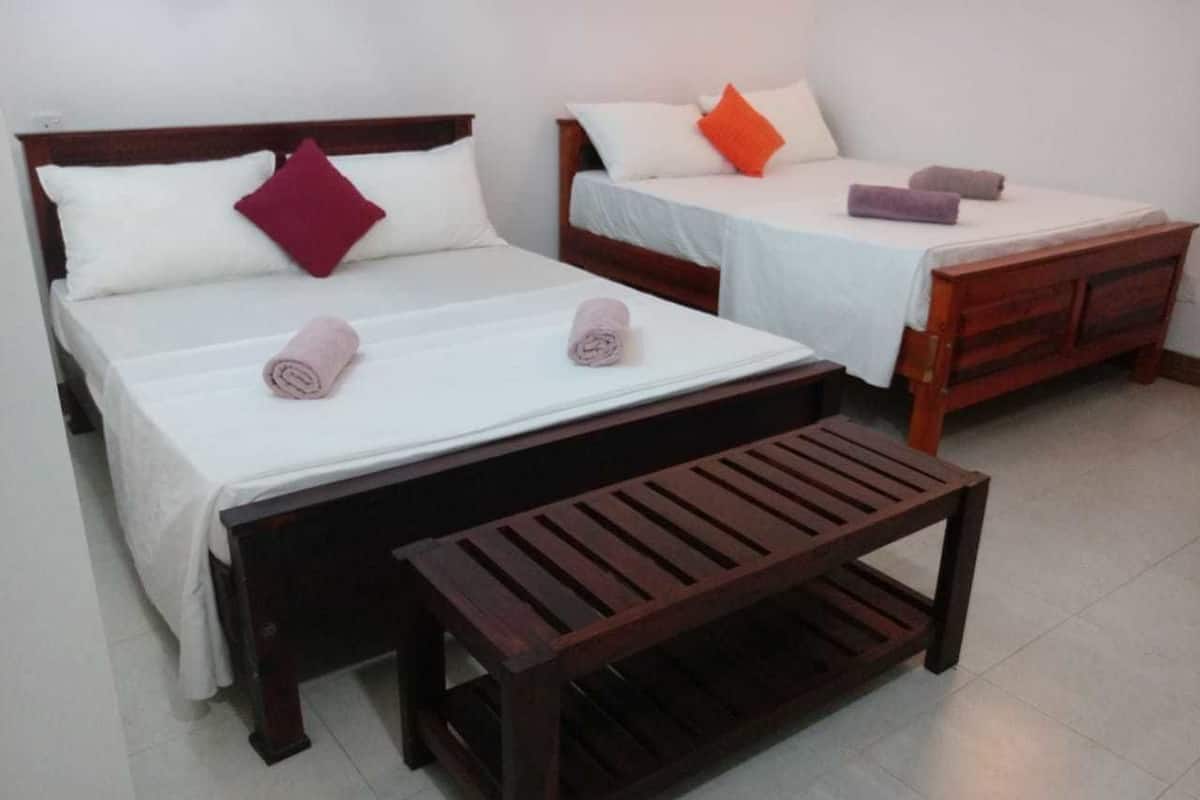Dos camas individuales con cabeceros de madera y ropa blanca, un banco de madera y un piso cubierto de baldosas blancas.