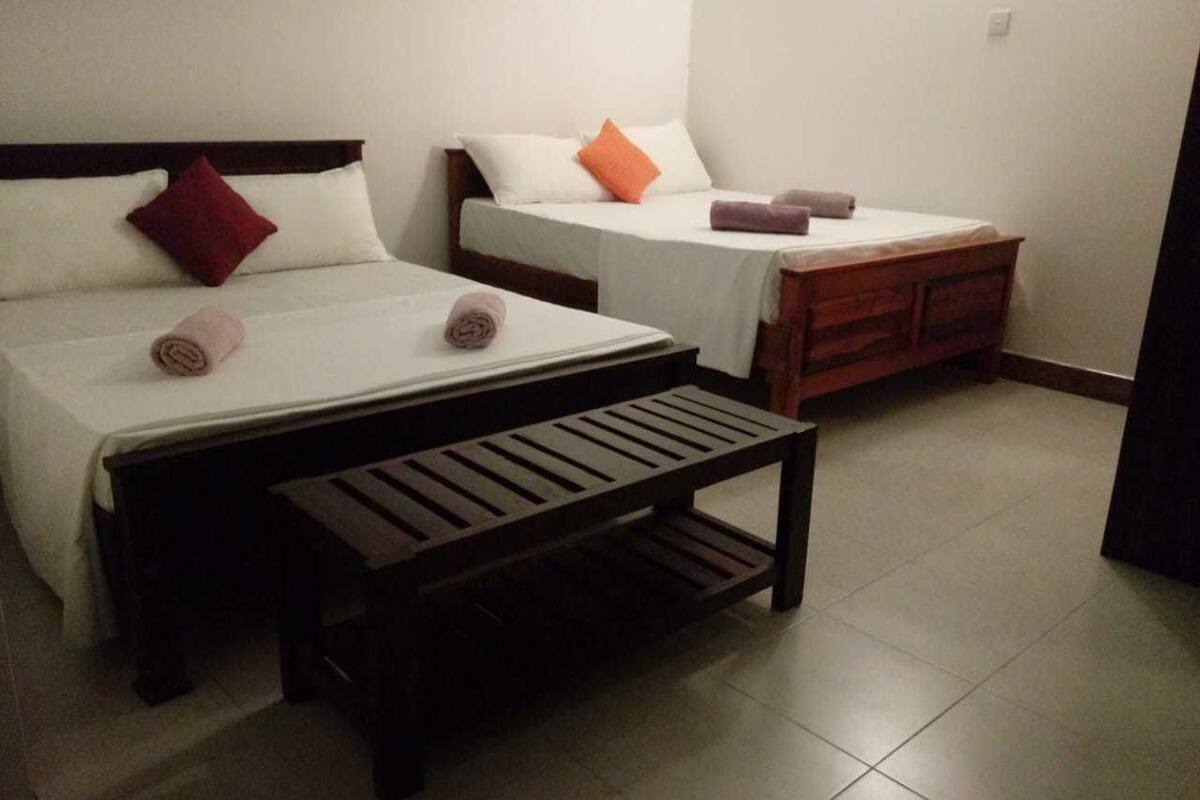 Habitación de hotel con dos camas individuales, cada una con una almohada y una toalla enrollada. Hay un banco de madera frente a las camas.
