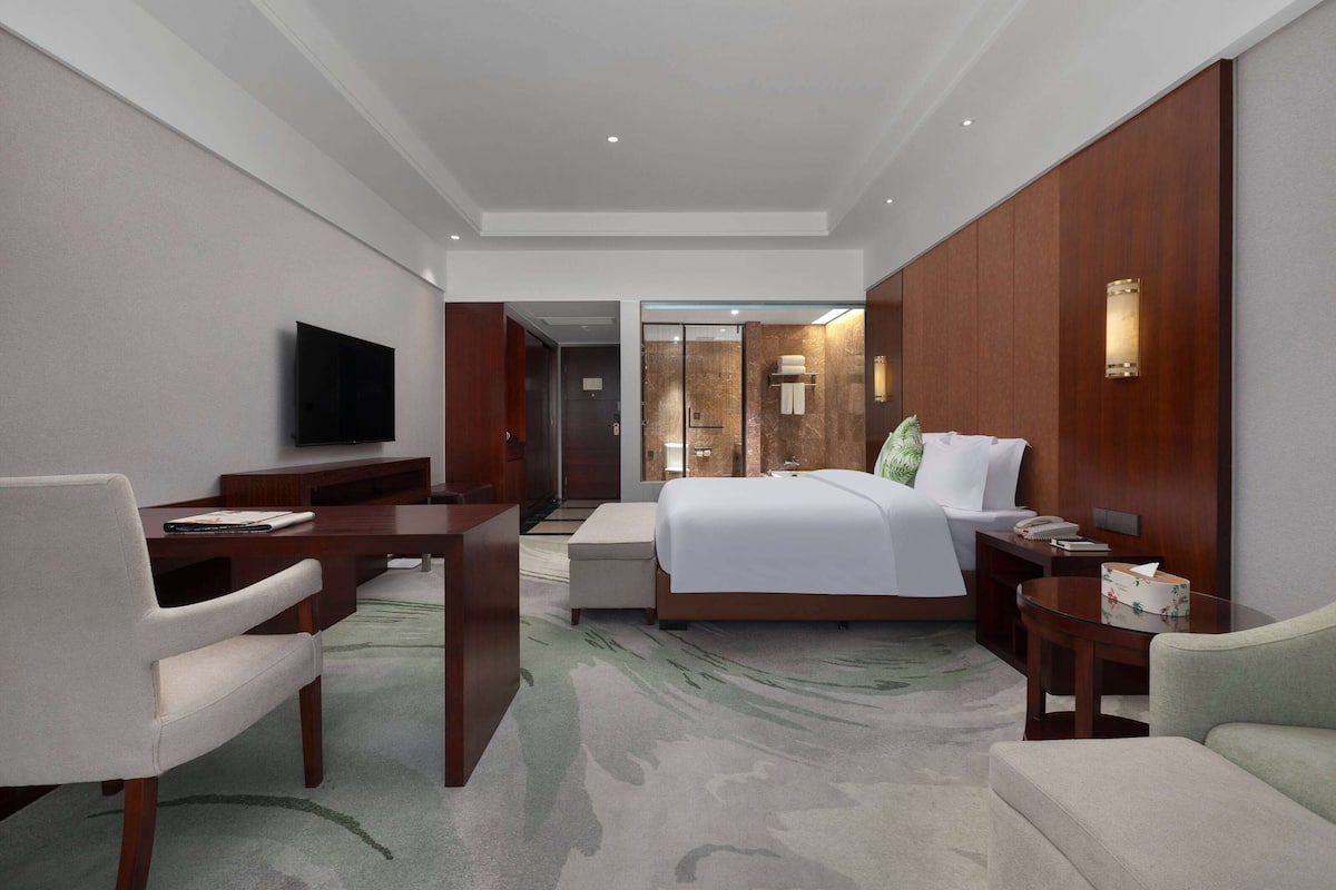 A hotel room with a bed, a desk, a chair, a television, and a bathroom visible through a glass door.