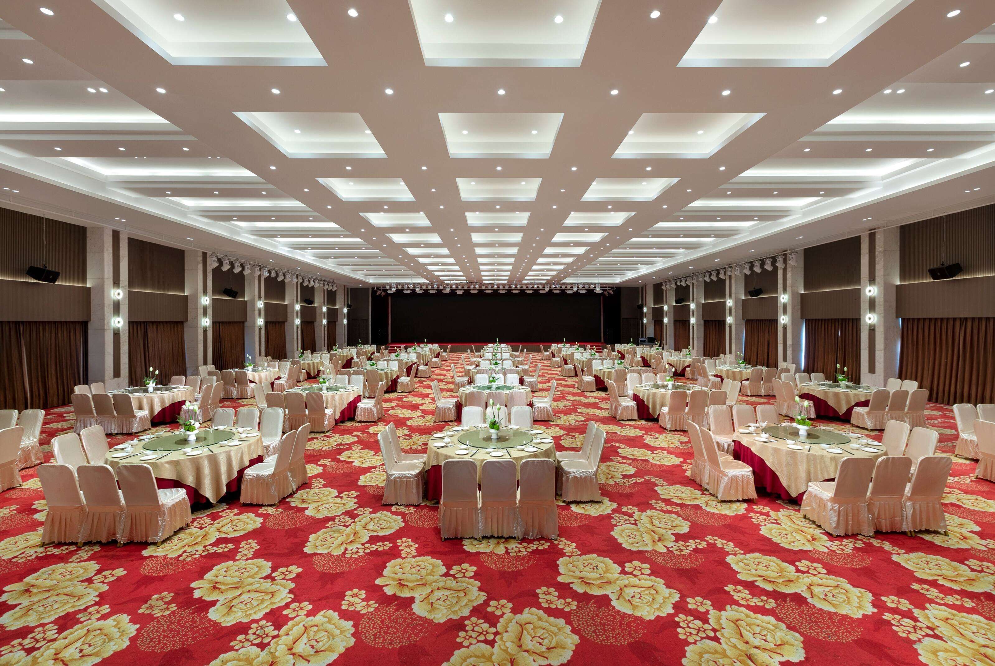 A banquet hall with round tables, white chairs, and a red floral carpet.