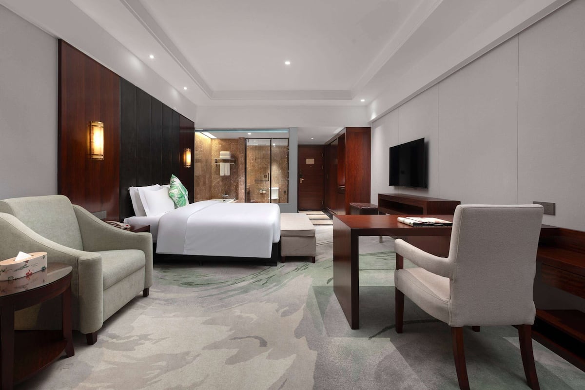 A hotel room with a bed, two armchairs, a desk, a television, and a bathroom visible through a glass door.
