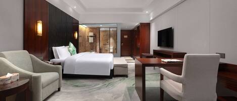 A hotel room with a bed, two armchairs, a desk, a television, and a bathroom visible through a glass door.