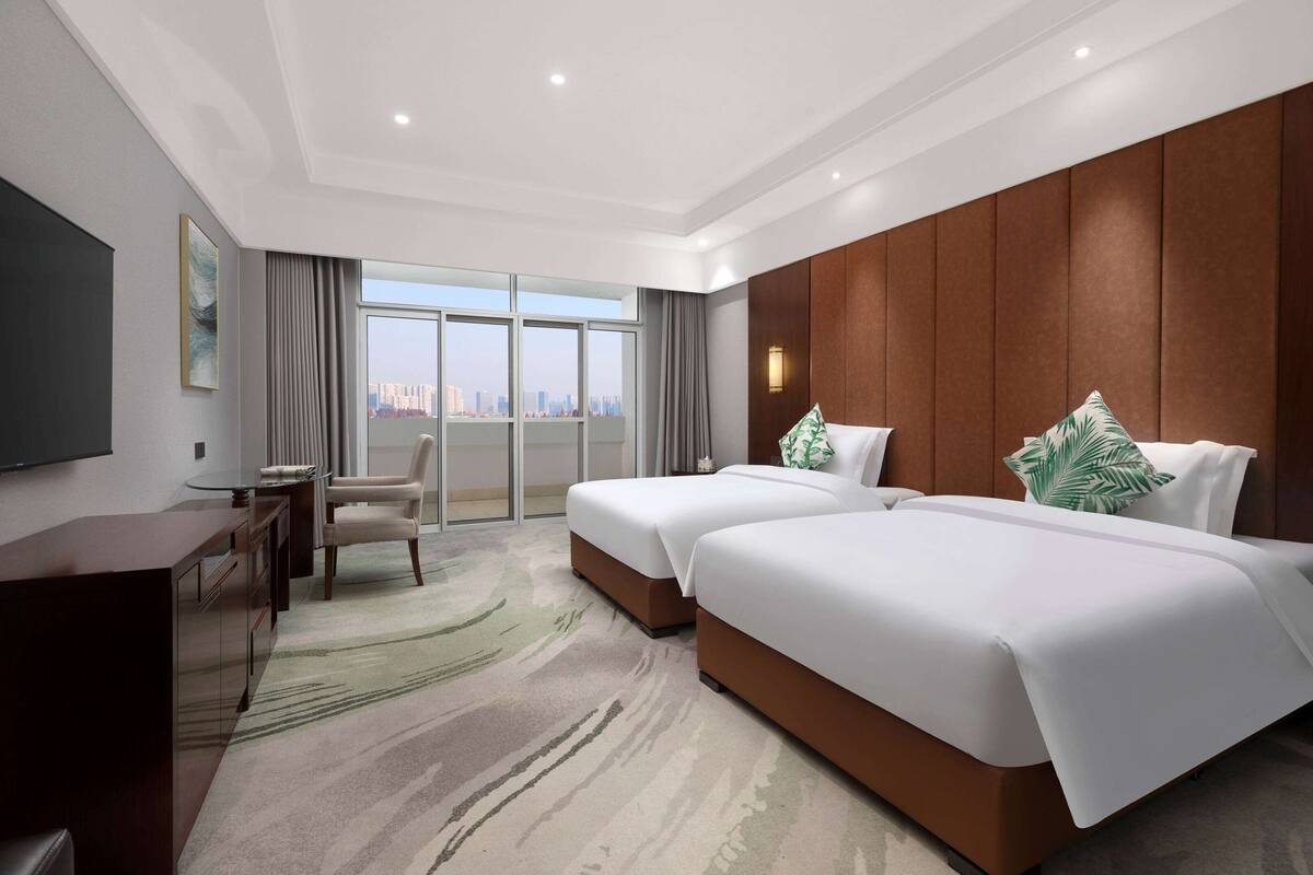 A hotel room with two beds, a desk, a chair, a TV, and a large window with a city view.