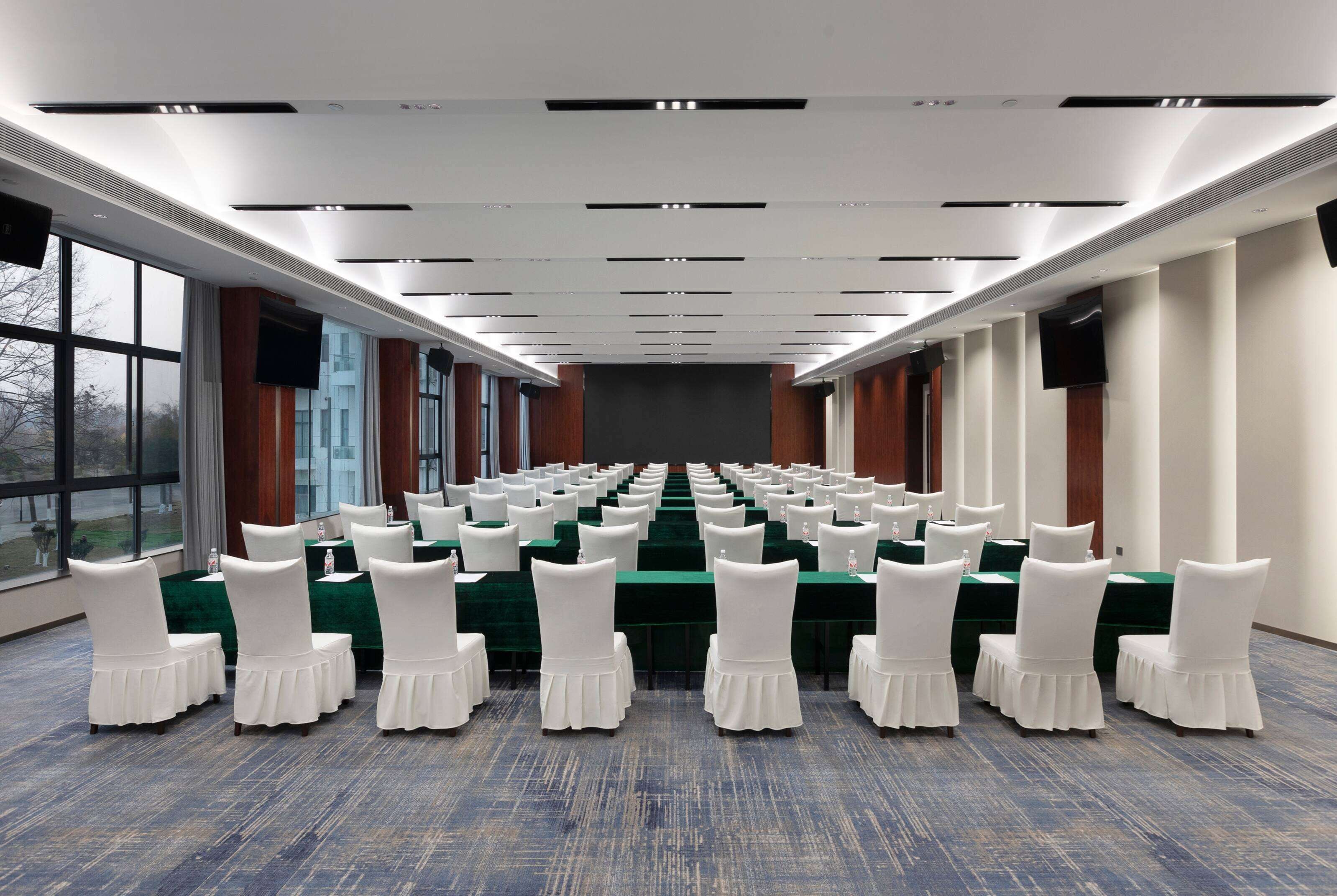 A conference room with rows of tables and chairs, large windows, and a flat-screen TV on the wall.