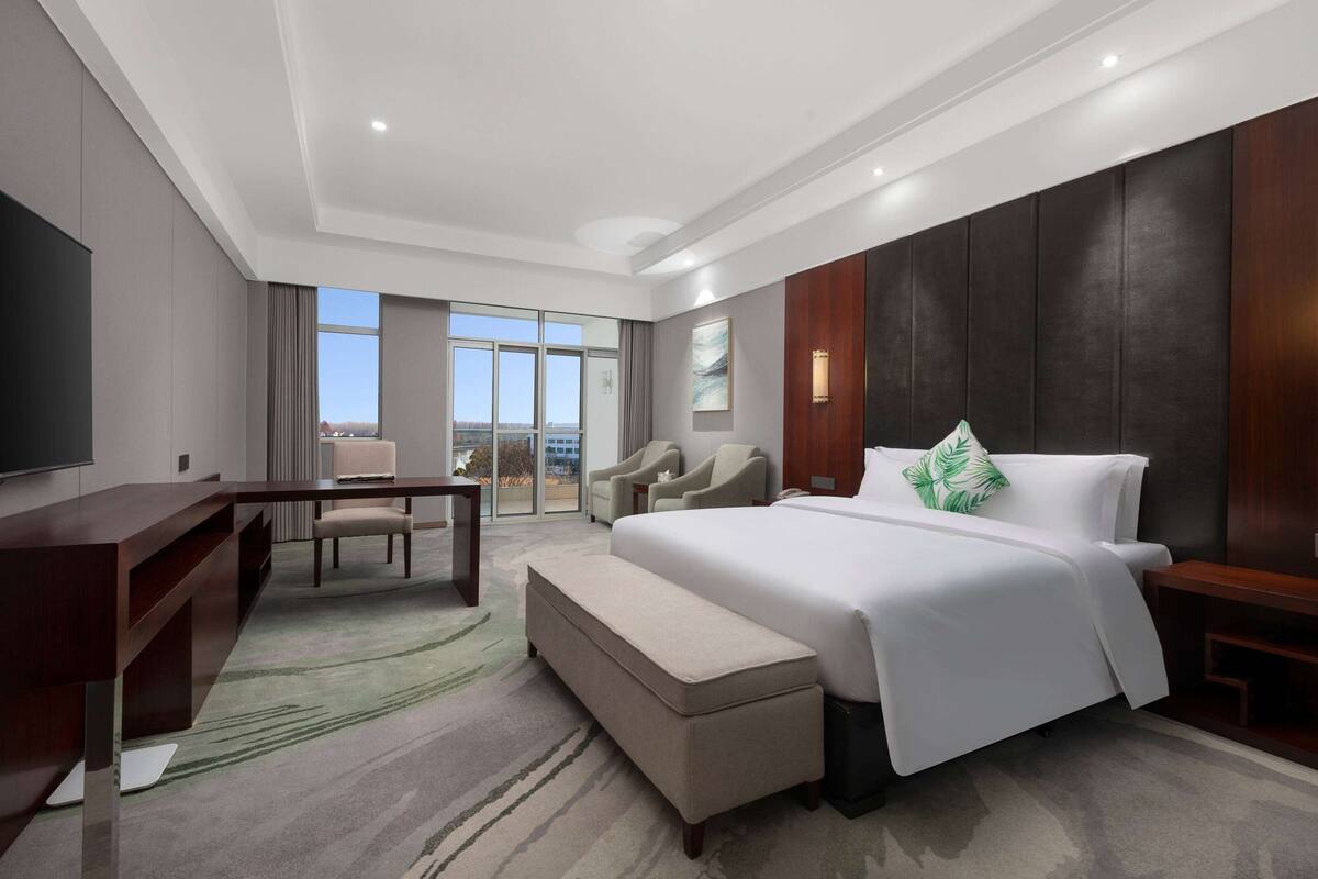 A modern hotel room with a large bed, a desk, a chair, and a view of the city.