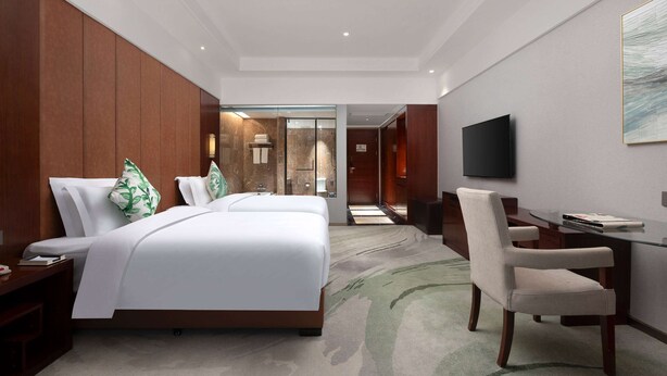Une chambre d’hôtel avec un grand lit, un bureau, une chaise, une télévision et une salle de bain visible à travers une porte en verre.