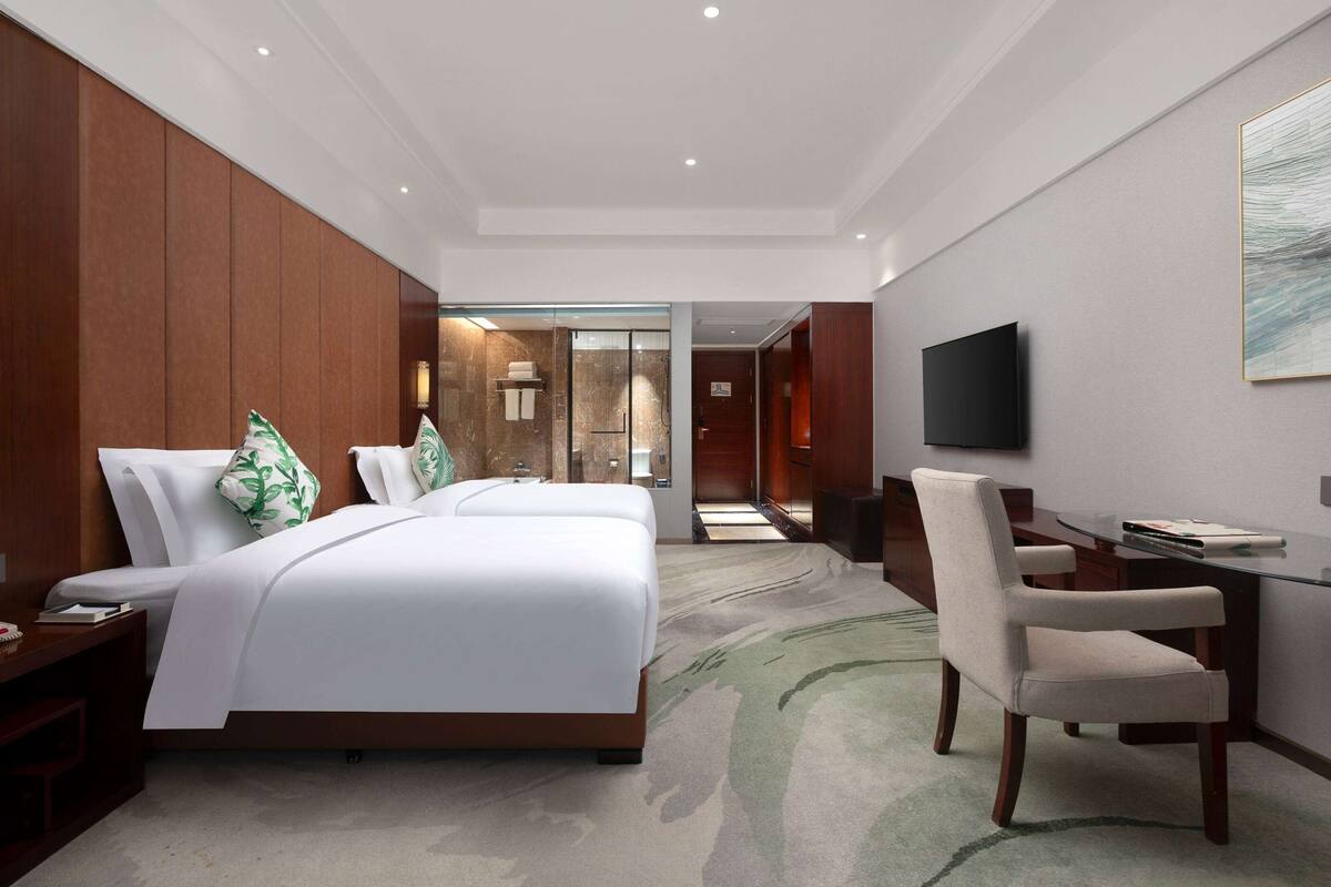 A hotel room with a large bed, a desk, a chair, a TV, and a bathroom visible through a glass door.