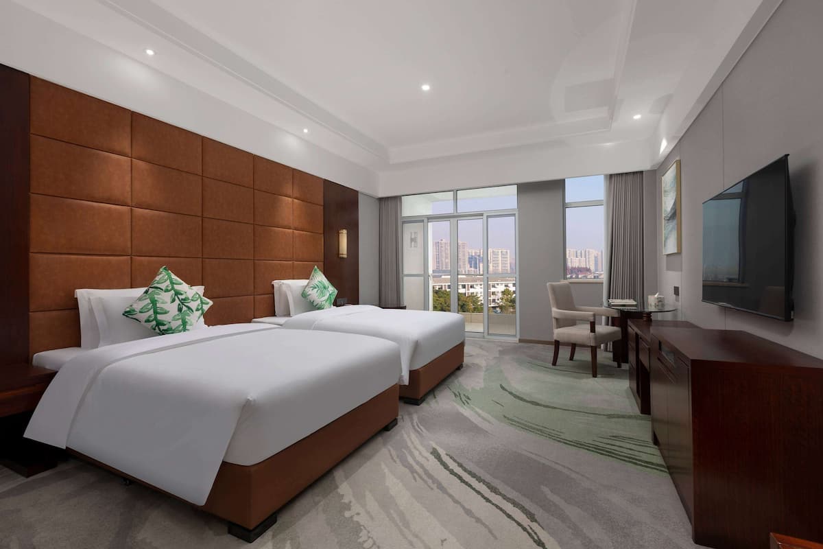 A hotel room with two beds, a desk, a chair, a TV, and a large window with a city view.