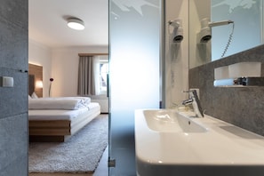 A modern hotel room with a bed, a large window, a bathroom with a sink and mirror, and a shower.