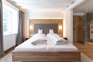 A modern bedroom with a wooden headboard, two bedside lamps, a large window with curtains, and a wooden wardrobe.