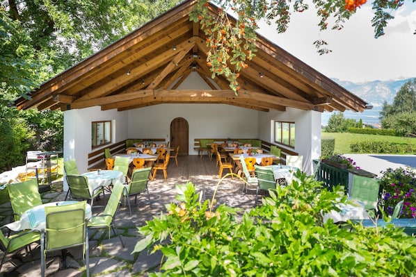 A covered outdoor dining area with tables and chairs, surrounded by greenery and a mountainous backdrop.
