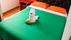 Ein Bett mit einer grünen Tagesdecke, zwei orangefarbenen Kissen und einem weißen Stoffhasen mit Äpfeln und einer Karte.