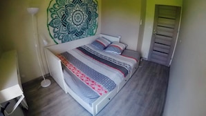 A bedroom with a bed, a floor lamp, a door, and a wall tapestry. - Charming and cosy rooms (Reze)