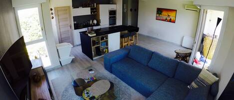 Uma sala de estar moderna com um sofá seccional azul, uma mesa de centro e uma TV em um suporte. Há uma área de cozinha com bancada e armários pretos.