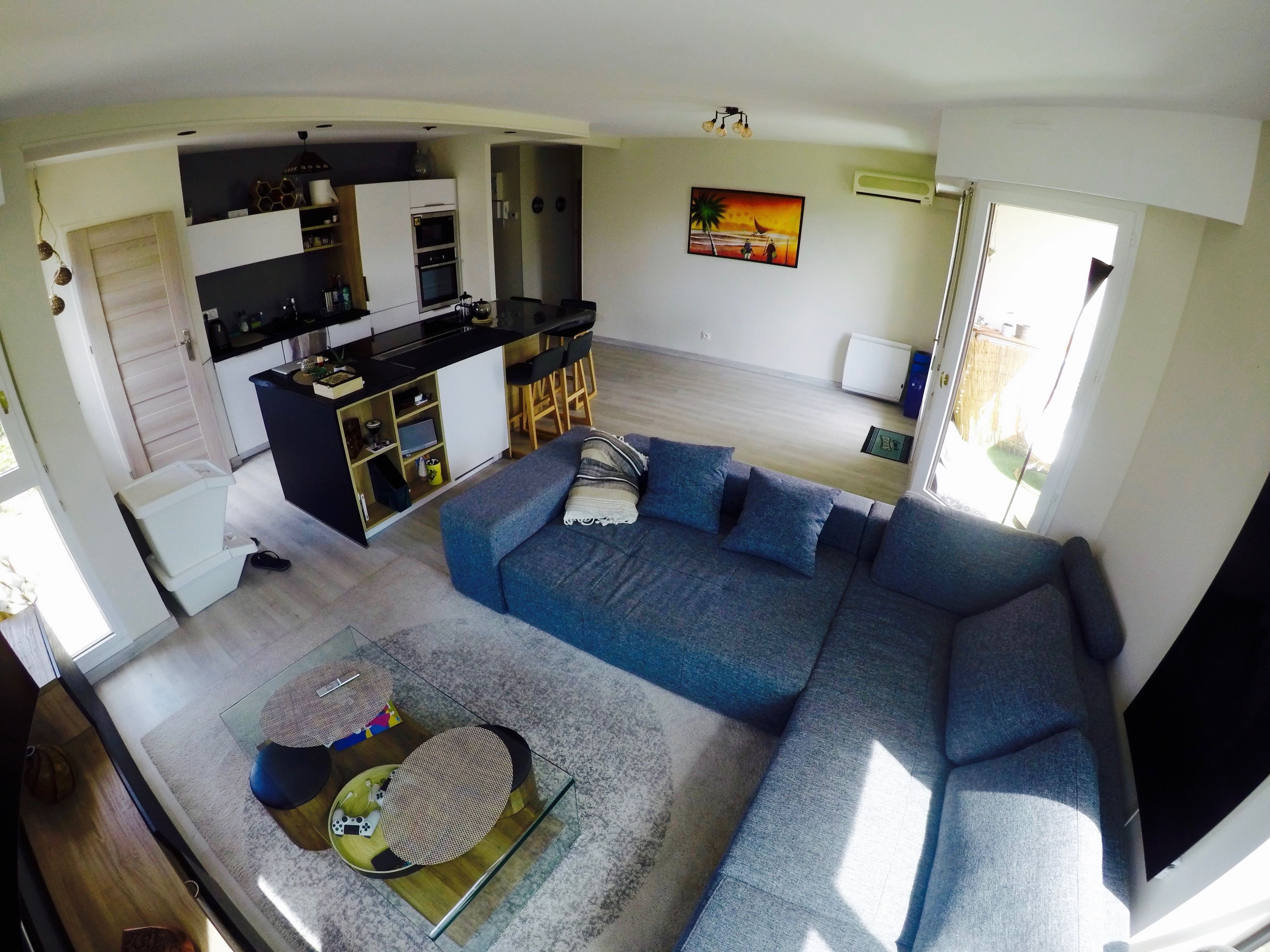 A modern living room with a large blue sectional sofa, a glass coffee table, and a kitchen area with a black countertop and white cabinets.