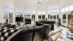 A spacious living room with a large black leather sofa, a glass coffee table, and a flat-screen TV. There are multiple windows providing natural light and views of the outdoors.