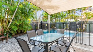 Um pátio externo com mesa e cadeiras de vidro, cercado por uma piscina e plantas tropicais.