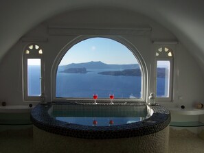 A room with a large arched window overlooking a body of water and distant land, a small pool with two red glasses, and a stone-tiled edge. - Caldera View Private Villa with Private Pool / Heated Jacuzzi / Amazing Sunset (Thira)