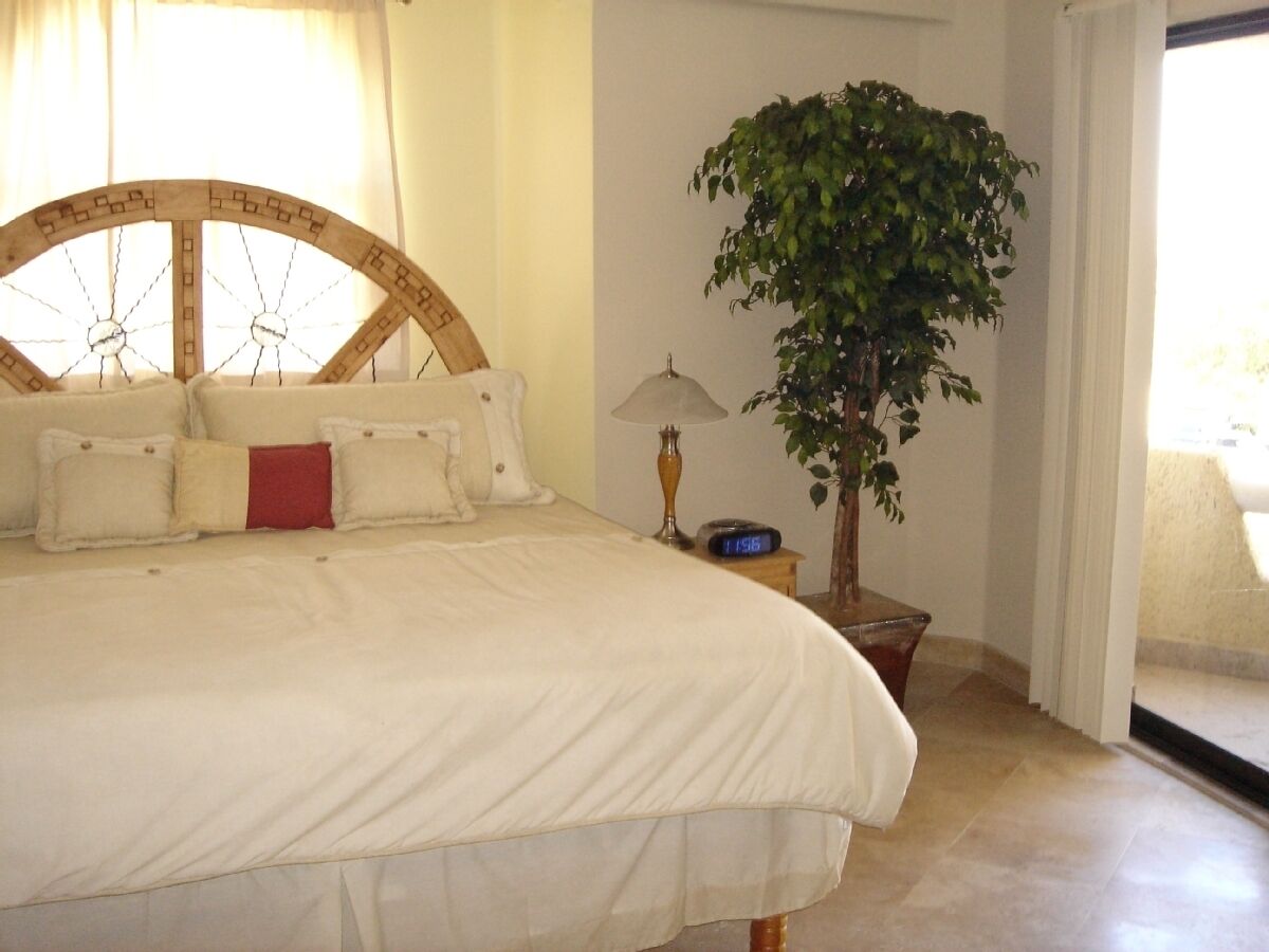 A bedroom with a large bed, a wooden headboard, a potted plant, a bedside table with a lamp, and a digital clock.
