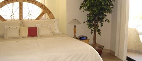 A bedroom with a large bed, a wooden headboard, a potted plant, a bedside table with a lamp, and a digital clock.
