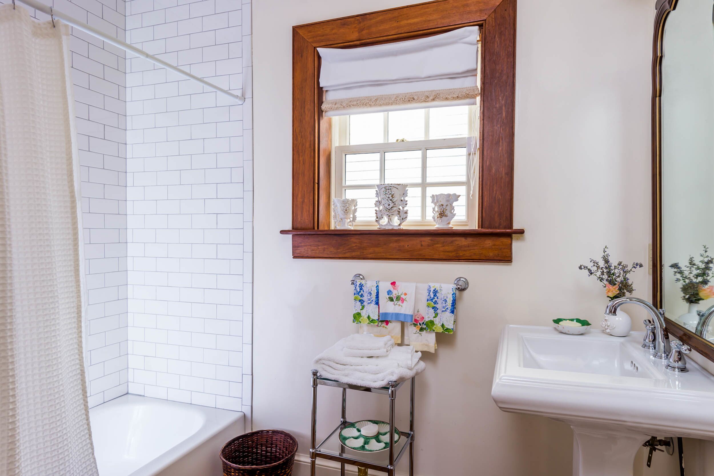 A bathroom with a white bathtub, a sink, and a mirror. There is a window with a wooden frame and a towel rack with towels.