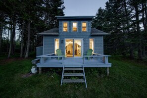 Une maison à deux étages avec une terrasse et deux chaises, entourée d’arbres.