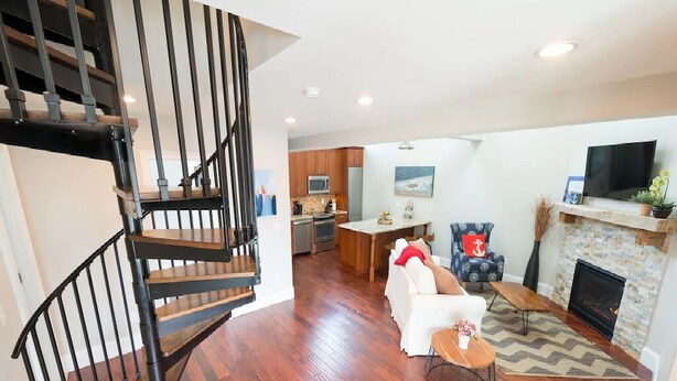 A modern living room with a staircase, a fireplace, and a kitchen in the background. - 2 Bedrooms w/Loft, Deck  and  Fireplace on Thames St (Newport)