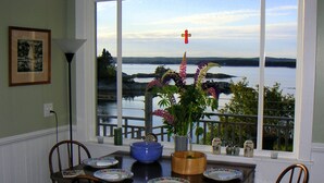 Uma área de refeições com uma mesa posta para quatro pessoas, uma janela com vista para um lago e colinas, e uma cruz pendurada acima da janela.