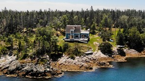 Uma casa moderna de dois andares com varanda, rodeada por uma floresta densa e um litoral rochoso.