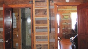 Interior de una cabaña de troncos con una escalera de madera que lleva a un dormitorio en el entrepiso, una chimenea de piedra y una sala de estar acogedora con un sofá.