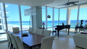 Ett rymligt vardagsrum med ett matbord, vita stolar, ett piano och utsikt över stranden.