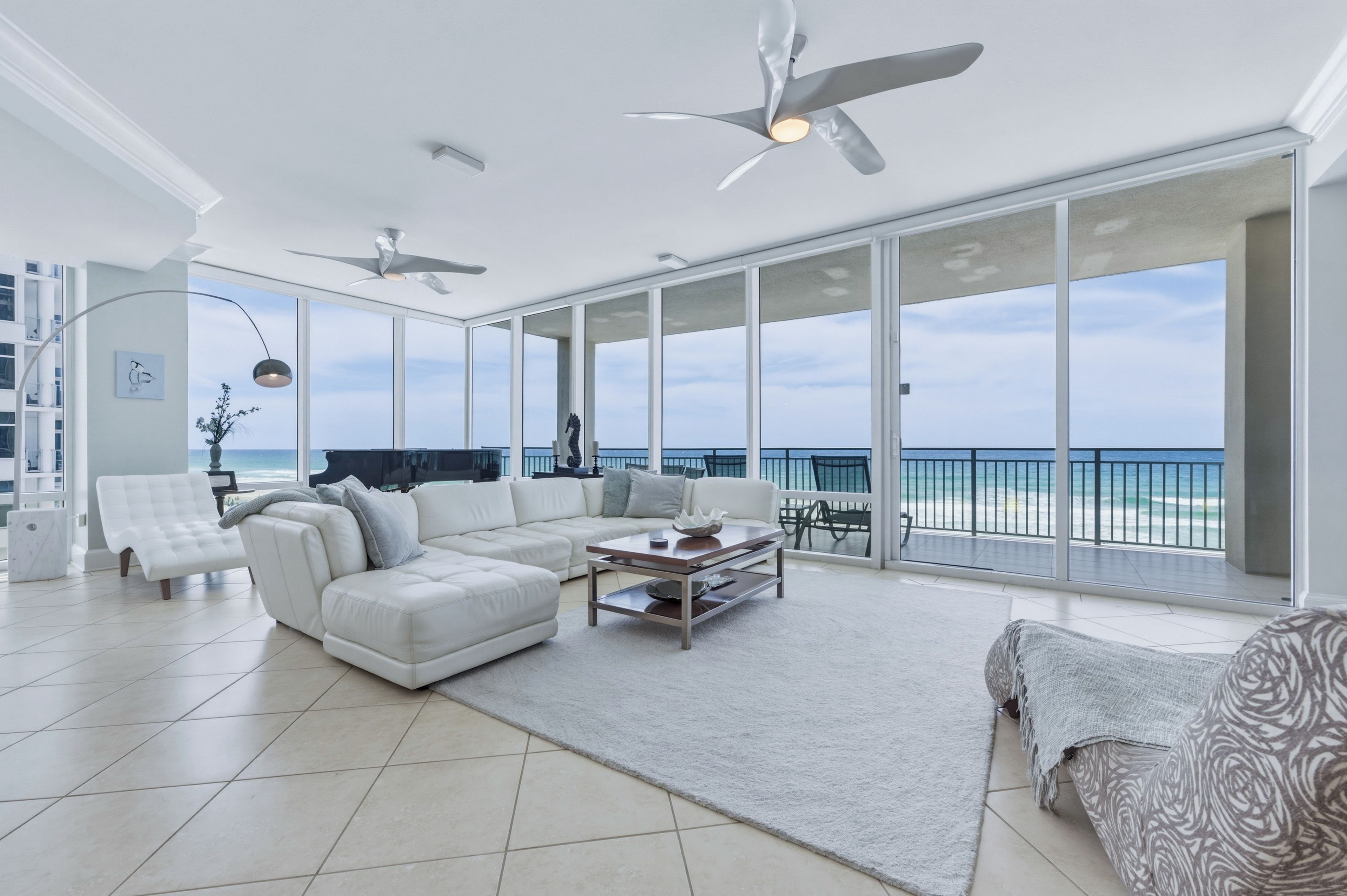 A spacious living room with a large white sectional sofa, a coffee table, and a view of the ocean through floor-to-ceiling windows.