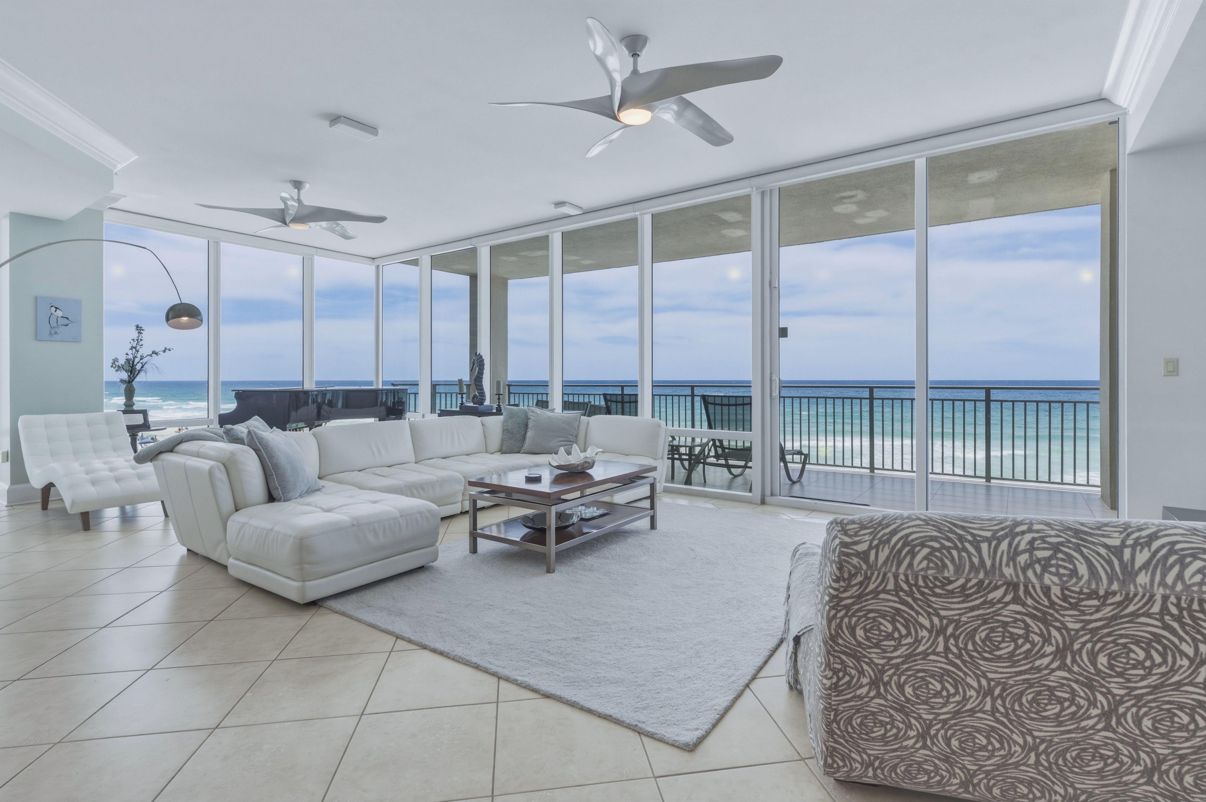 A spacious living room with a large window overlooking the ocean, a ceiling fan, and a modern white sofa.