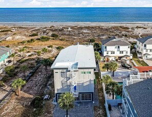 A coastal residential area with a modern two-story house featuring a flat roof, a central staircase, and a small front yard with palm trees. - Private Heated Pool, Beach Access, All King Beds (Tybee Island)