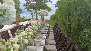Una larga mesa de comedor dispuesta con flores blancas, velas y platos, con vista a una playa.