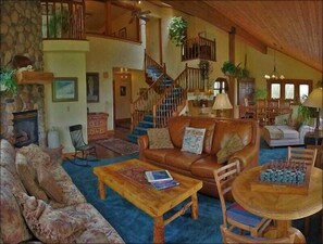 A spacious living room with a stone fireplace, a wooden staircase, and a cozy seating area with a brown leather sofa and patterned armchairs.
