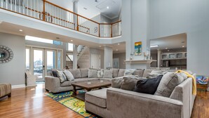 A spacious living room with a large sectional sofa, a coffee table, and a colorful rug. There is a staircase leading to an upper level, and a kitchen area is visible in the background.