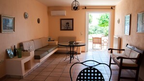 A living room with a sofa, a round table, and a chair. There is a door leading to an outdoor area with a chair and a table. - Villa Hera At Cape Sounion (Lavreotiki)