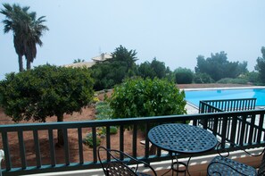 A poolside area with a table and chairs, surrounded by greenery and a view of a building in the background. - Villa Hera At Cape Sounion (Lavreotiki)