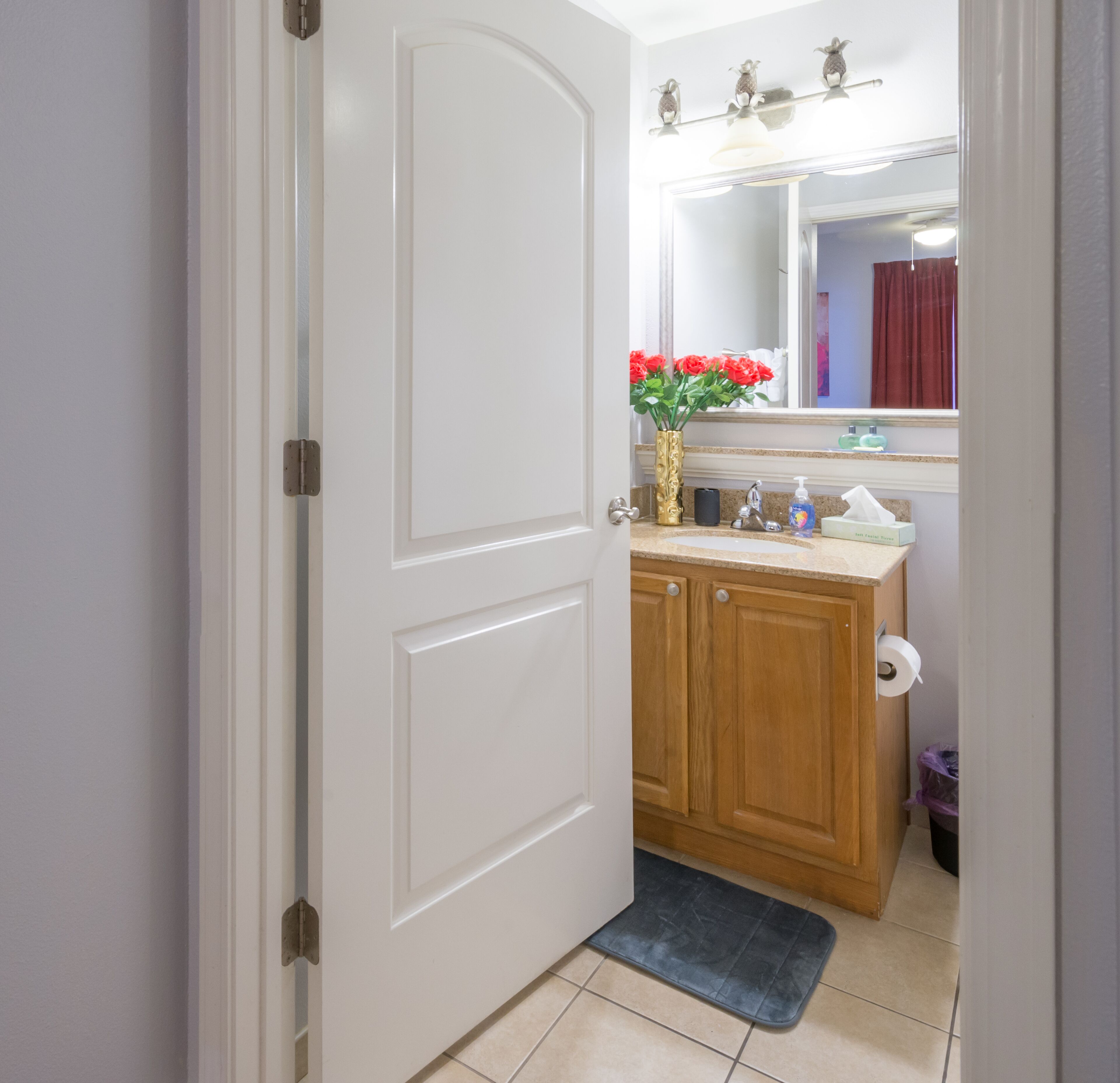 A bathroom with a white door, wooden cabinets, a sink with a mirror above, and a vase with red flowers.